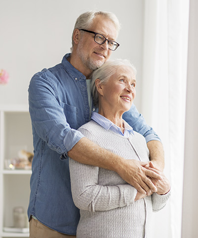 Ventajas para Particulares - Una pareja senior abrazados mirando por la ventana de su casa sonriendo