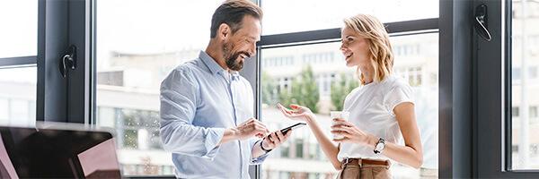 Carteras Premium - Hombre con camisa y móvil en la mano, y mujer de negocios con cafe en la mano sonriendo en la oficina Carteras Premium - Hombre con camisa y móvil en la mano, y mujer de negocios con cafe en la mano sonriendo en la oficina